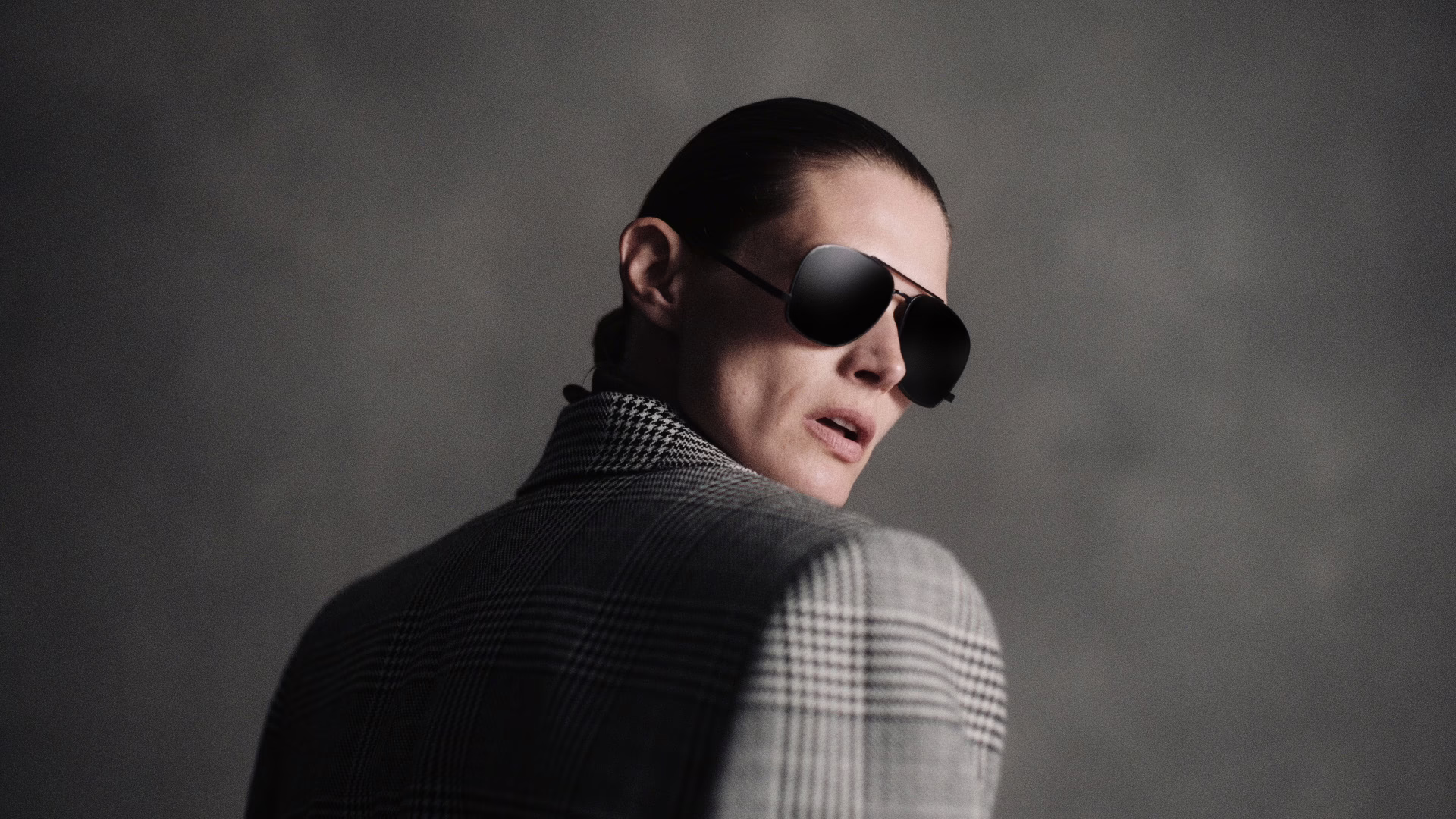 Black and white portrait of a model in aviator sunglasses looking back over a houndstooth blazer shoulder